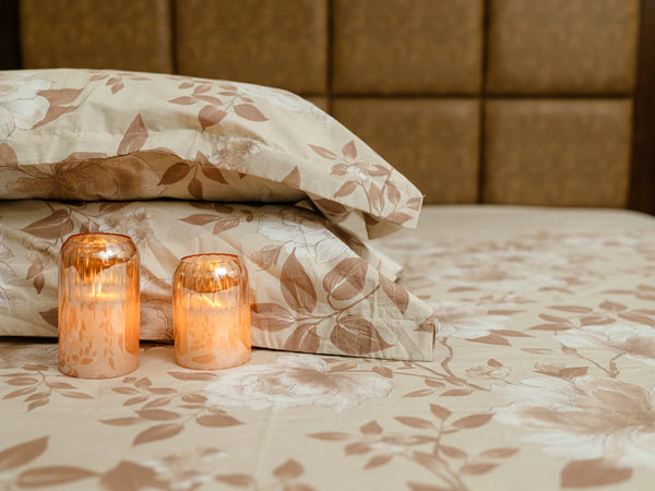 Two pillow covers stacked over one another with two decorative candles on a premium fawn and caramel floral 500 thread count organic cotton bedsheet with matching pillow covers styled on a luxury bed