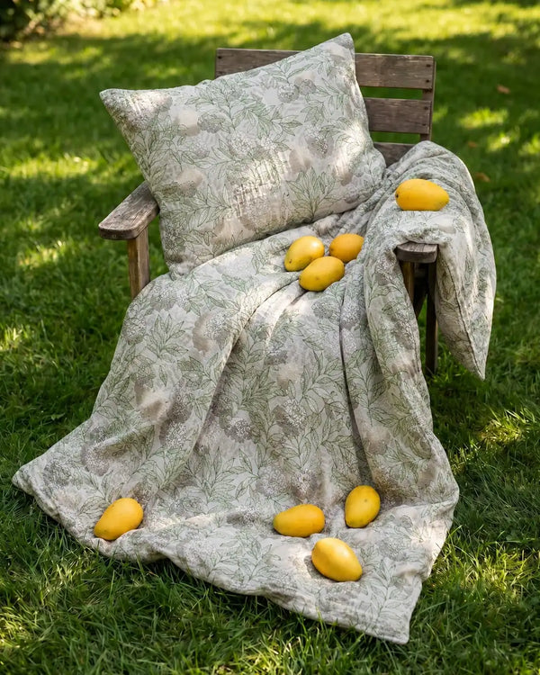 Alchemy luxury cotton bedsheet displayed outdoors with matching pillow cover, featuring earthy grey florals and vibrant green leaf motifs spreads on lifestyle wooden chair with mangoes over it