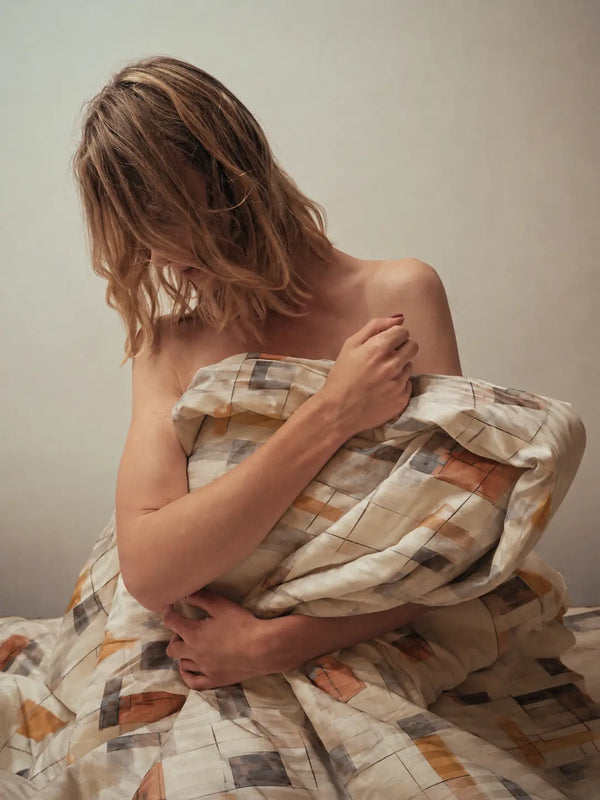 A lifestyle model is sitting on a bed wrapped with Spectrum luxury off white and mocha abstract cotton bedsheet styled in an elegant lifestyle shot highlighting soft premium bedding comfort.