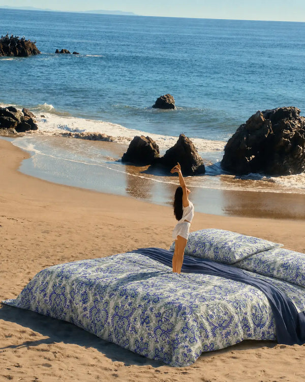 Kinetic luxury white and indigo cotton bedsheet styled in a beach setting wherein a lifestyle model is standing on the bed, showcasing blue damask design and serene premium comfort