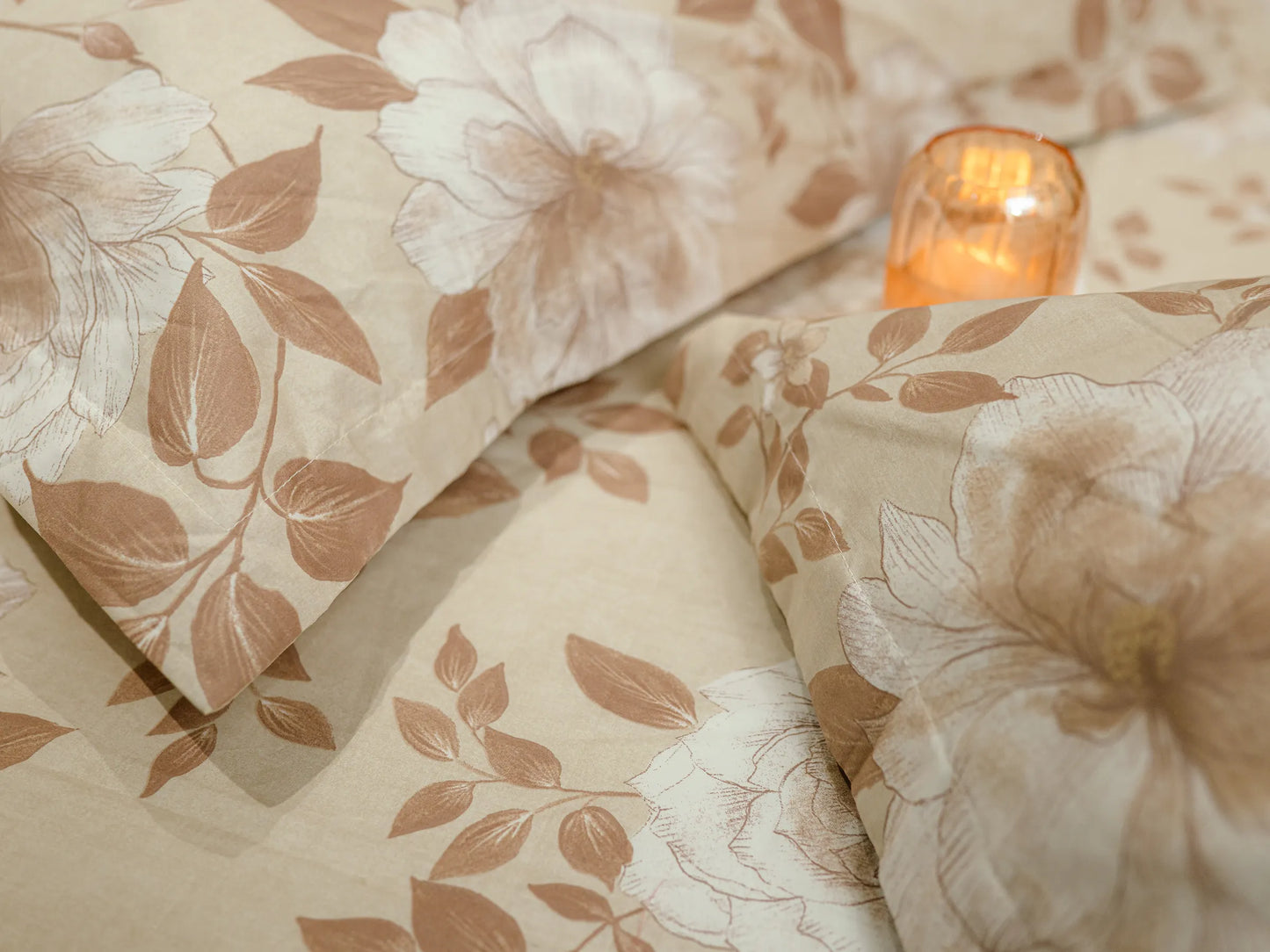 Corners of two pillow covers kept bedside a decorative candle kept on premium fawn and caramel floral organic cotton bedsheet highlighting premium 500 thread count fabric and floral detailing
