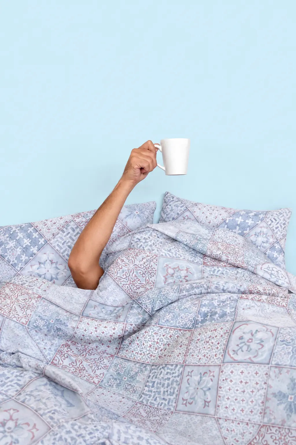 A lifestyle model holding a cup of coffee on Echelon indigo blue luxury bedsheet styled in a cozy morning scene with artistic abstract tile print and premium cotton finish