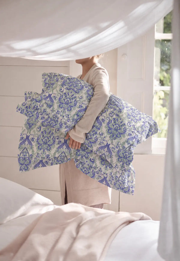 A lifestyle model is holding Kinetic white and indigo pillow cover held in a bright bedroom, highlighting elegant blue damask floral artwork and soft cotton finish